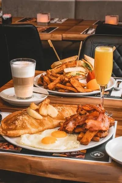 Abundant breakfast plates with eggs, bacon, toast, fries, sandwiches, a latte, and orange juice.