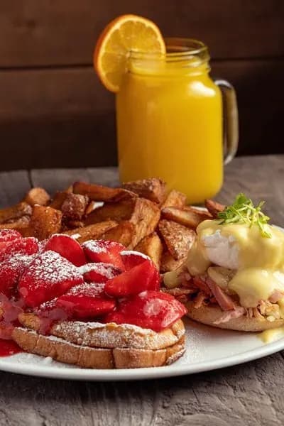 A hearty breakfast plate featuring strawberry French toast, eggs Benedict, fries, and orange juice.